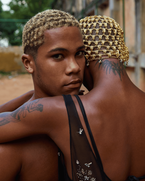 Dois jovens negros com cabelo descolorido e reflexo alinhado se abraçam em cenário de favela no Rio de Janeiro; imagem faz parte da campanha Sunset to Sunrise da Rabanne e representa a estética afetiva, orgulhosa e estilosa das periferias cariocas.